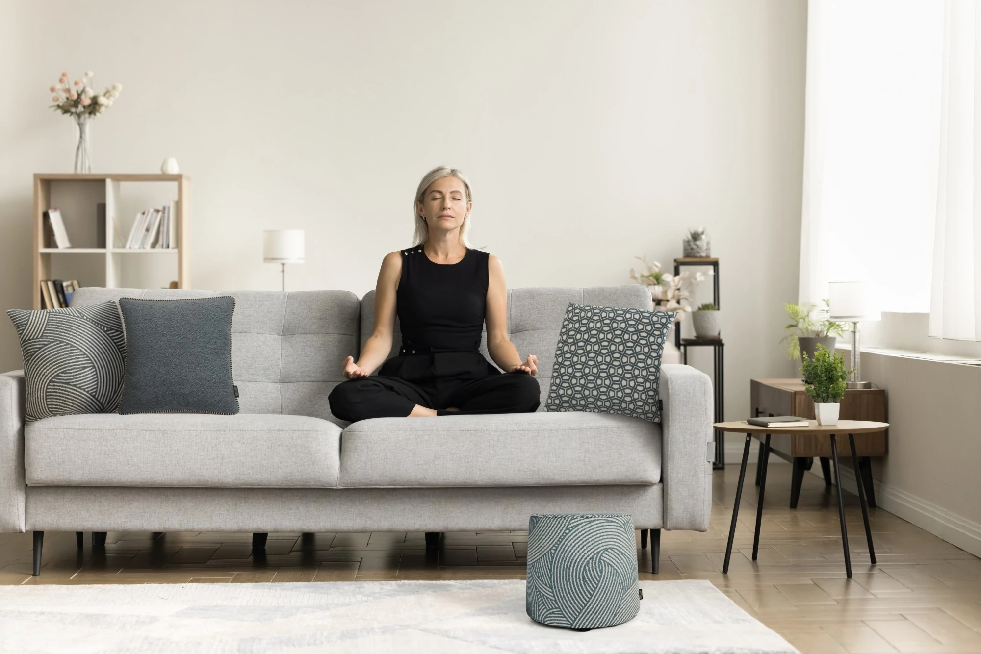 zenheader Frau meditiert auf einem Sofa mit Kissen und Pouf der Zen-Garden-Kollektion von Rohleder.