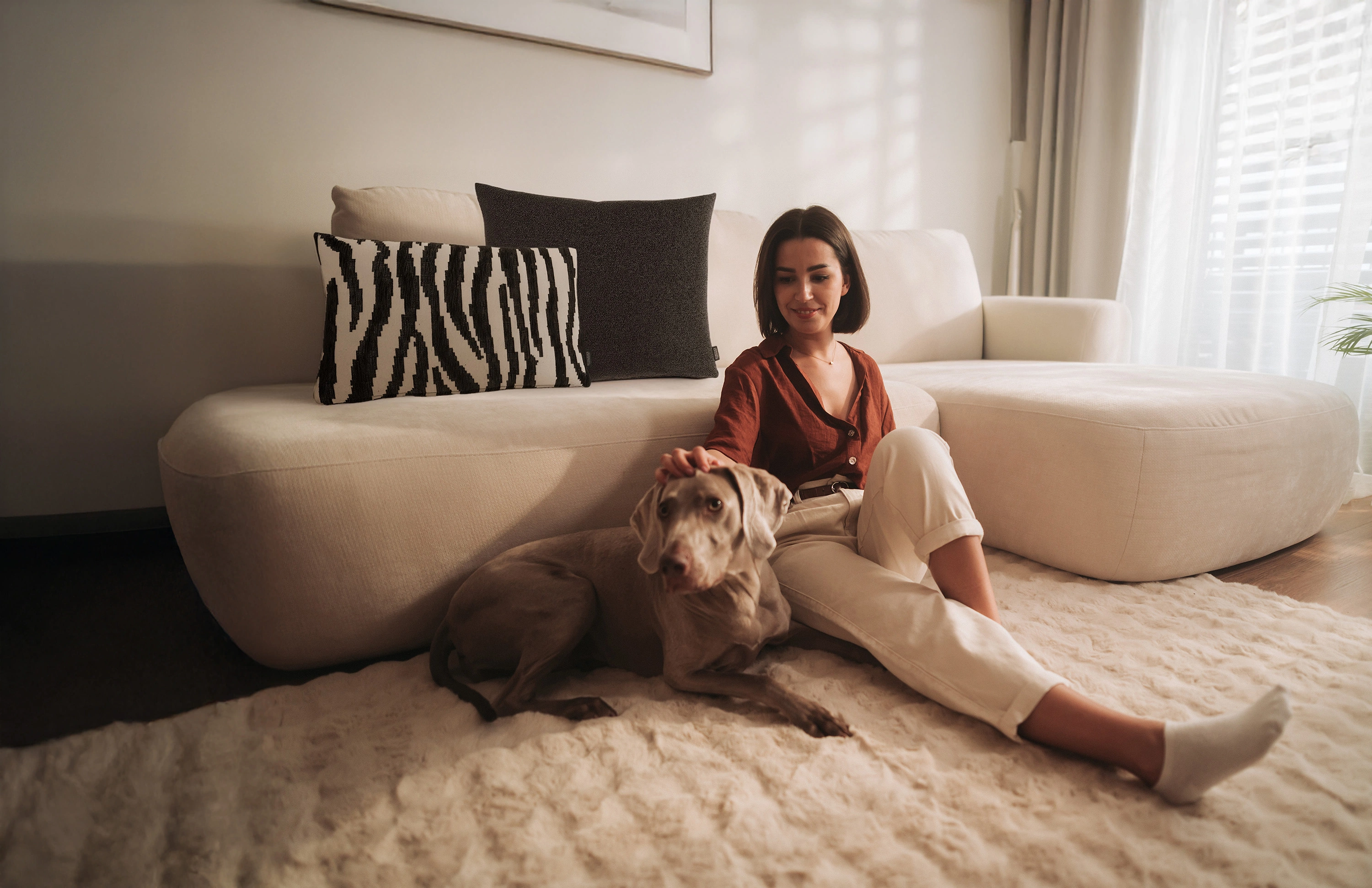 rohleder-savannah Frau sitzt auf hellem Sofa und streichelt Hund, auf dem Sofa liegen Zebra- und Ono-Kissen aus der Savannah Kollektion von Rohleder
